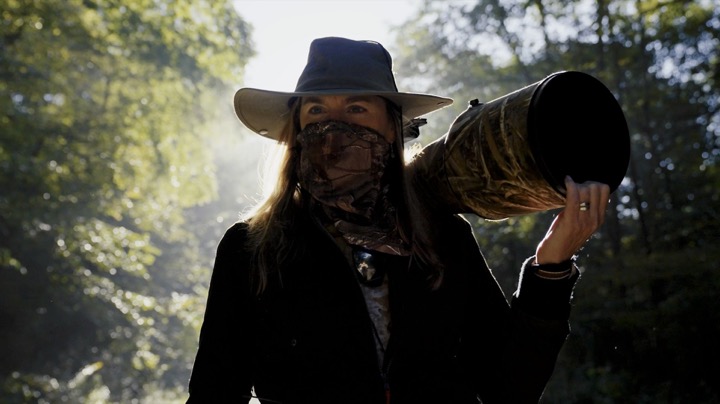 Photographer in a hat and face covering carrying a large telephoto lens in the woods